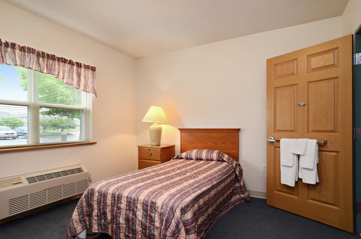 A cozy bedroom with a single bed, nightstand, and lamp beside a window.