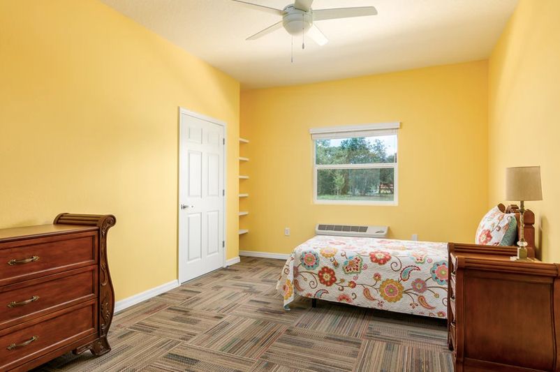A minimalist bedroom with yellow walls, featuring a single bed and a window, complemented by a ceiling fan and carpeted flooring.
