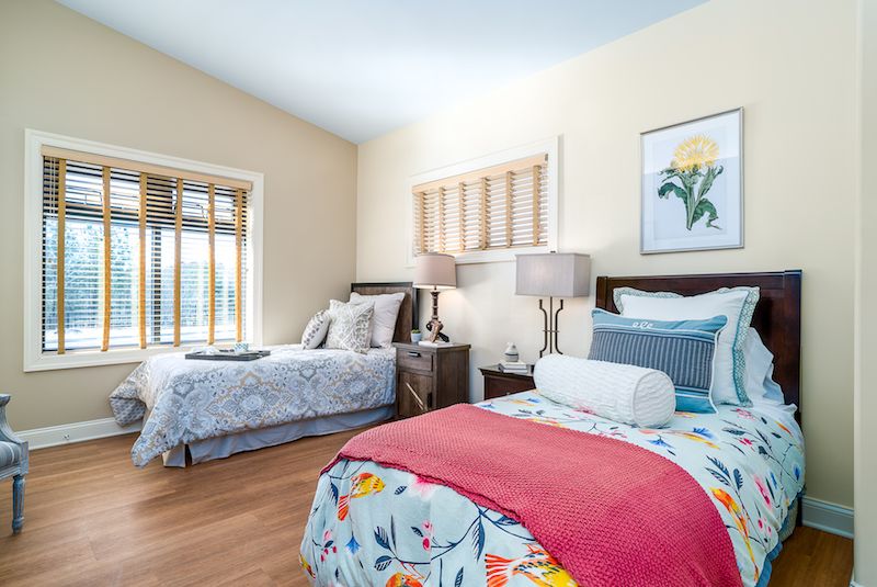 A well-lit bedroom featuring two neatly made beds with colorful bedding and decorative pillows, complemented by bedside tables and artwork on the walls.