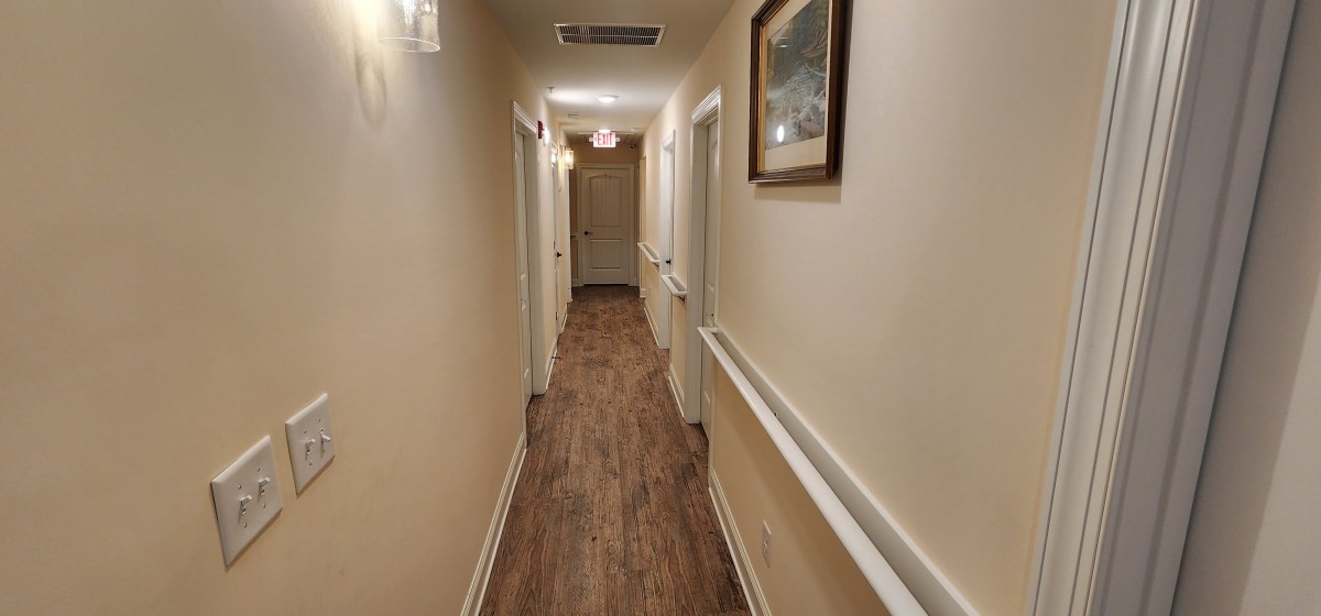 A well-lit indoor hallway with wooden flooring, lined with doors and a framed picture on the wall.