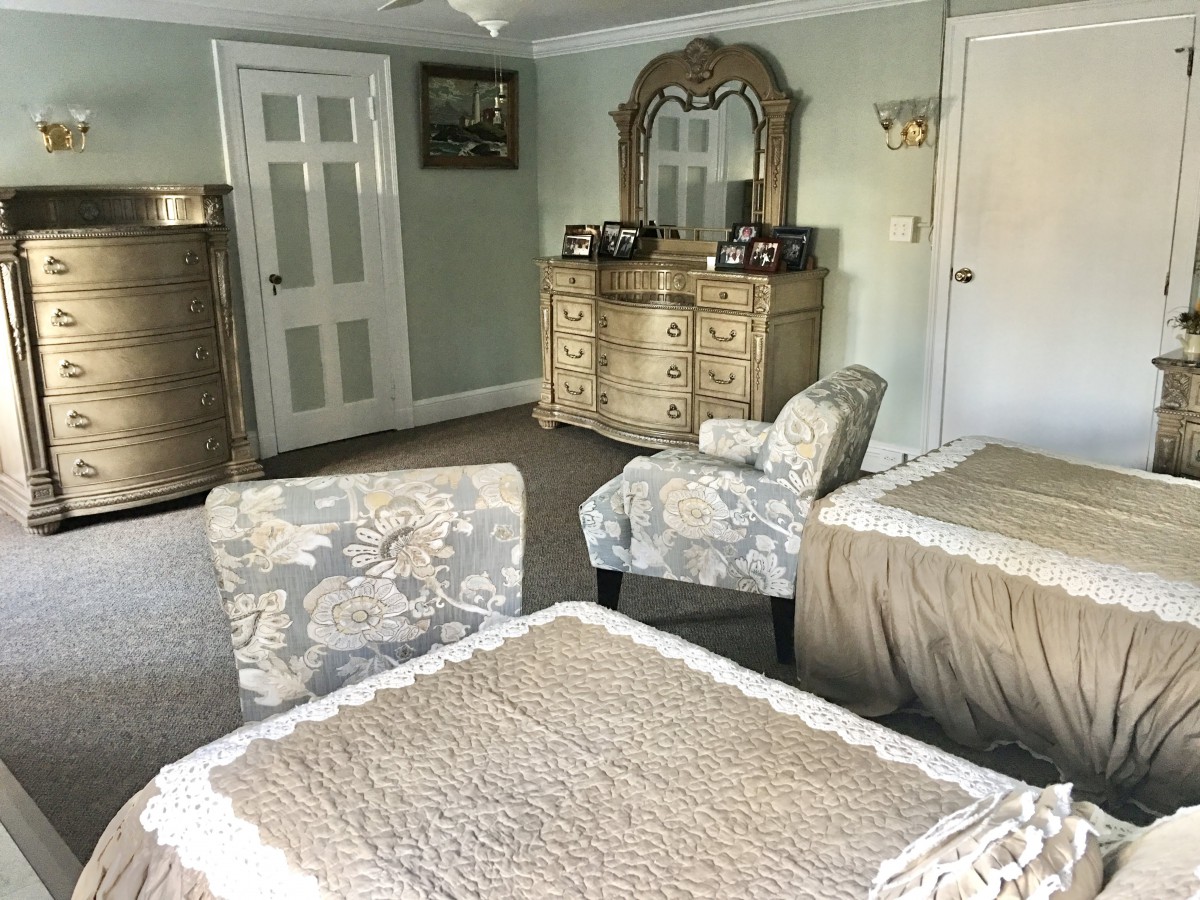 A cozy bedroom featuring a dresser with a mirror, upholstered chairs, and a table with lace covering.
