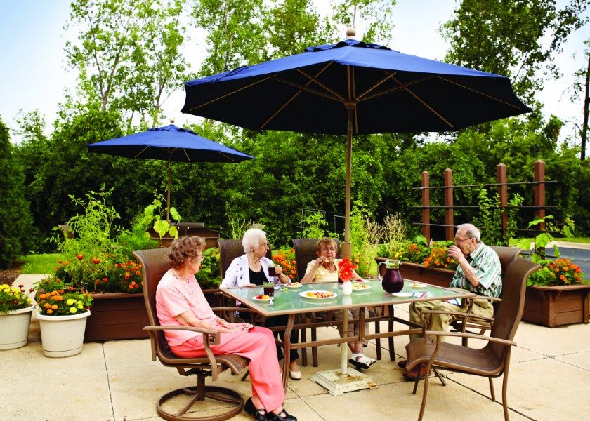 A group of seniors enjoying a relaxed afternoon on an outdoor patio, surrounded by greenery and shaded by blue umbrellas.