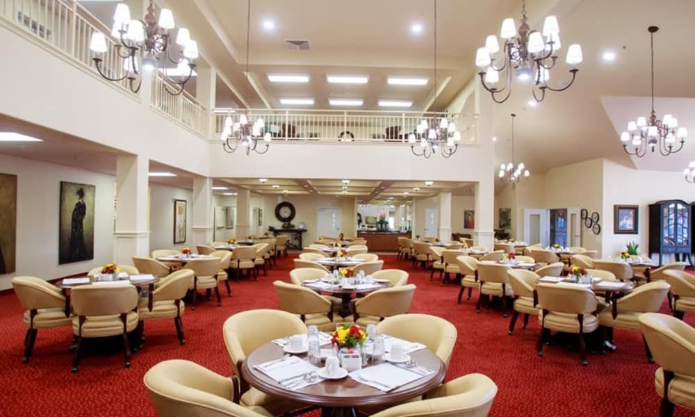 An elegant dining room with chandeliers, set with tables and chairs on a red carpet.