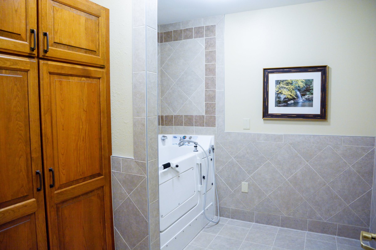 A bathroom featuring a walk-in tub, tiled walls, and a wooden cabinet with a framed picture on the wall.