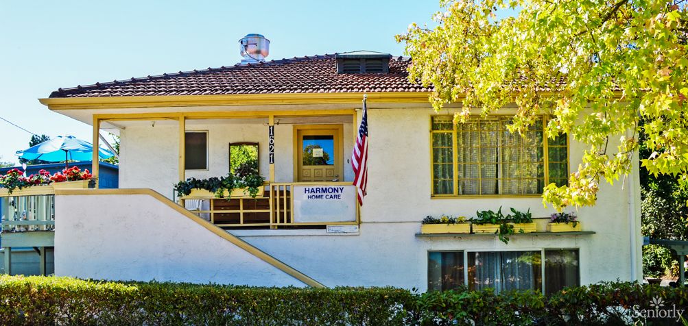 A two-story house with yellow trim and an American flag displayed, surrounded by trees and bushes.