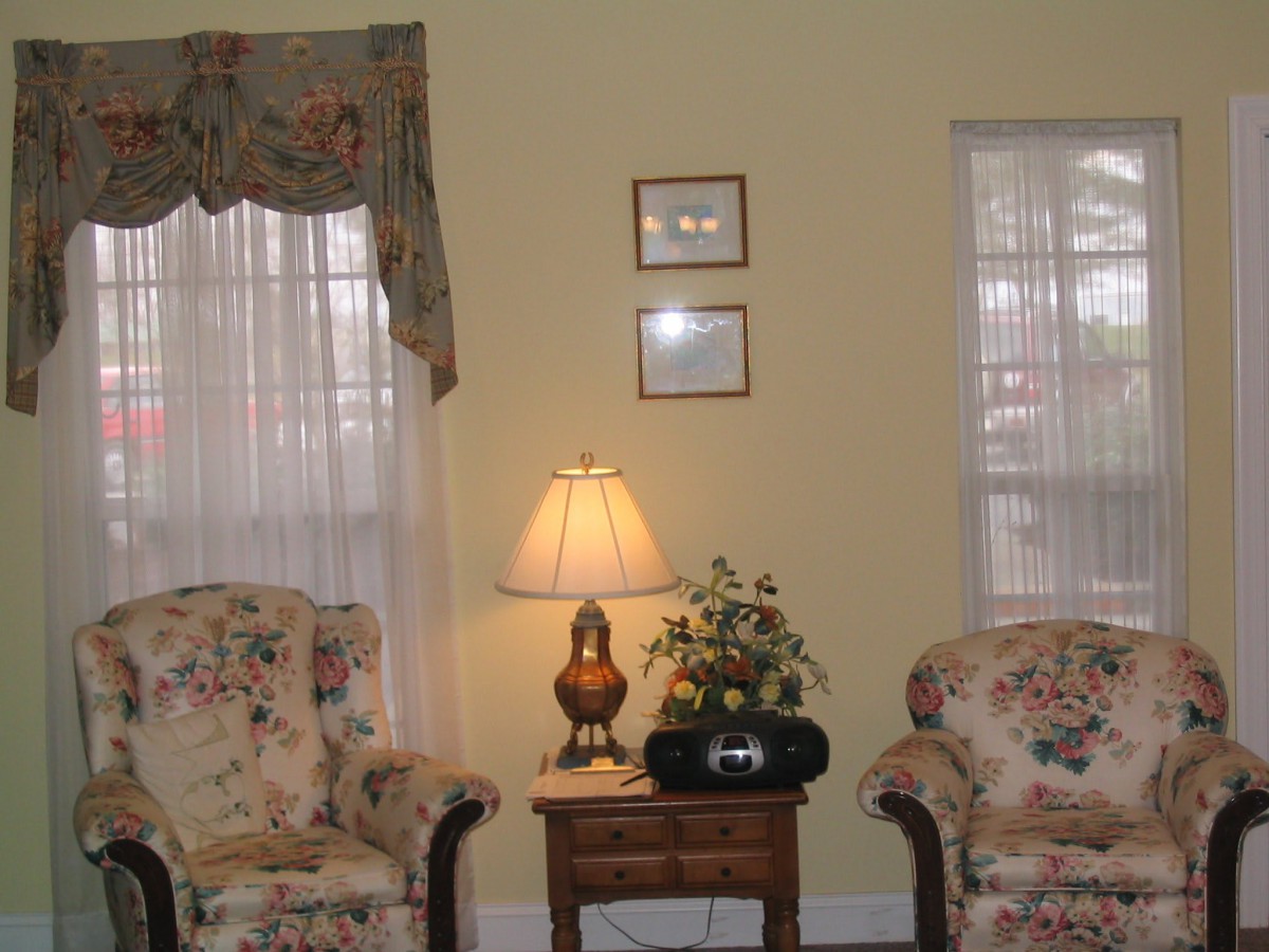 A cozy living room setup with two armchairs, a side table holding a lamp and plant, and decorated with sheer curtains.