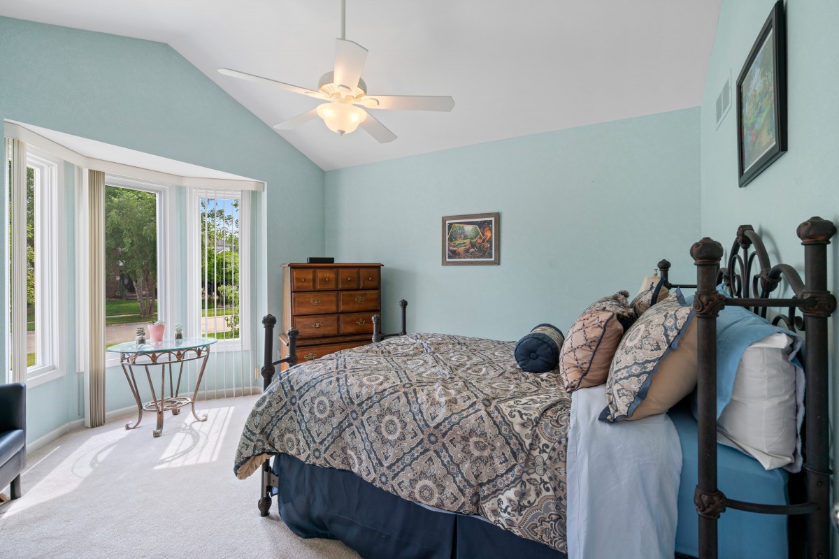 A cozy bedroom with a patterned bedspread, decorated in soothing colors and illuminated by natural light from a large window.