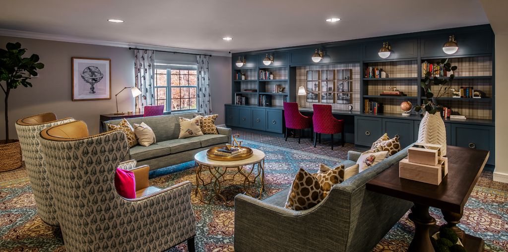 A cozy living room featuring comfortable sofas, a patterned rug, bookshelves, and a desk area with red chairs.