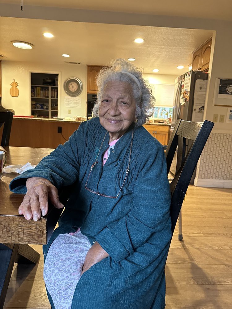 An elderly woman smiling while sitting at a table in a cozy kitchen.