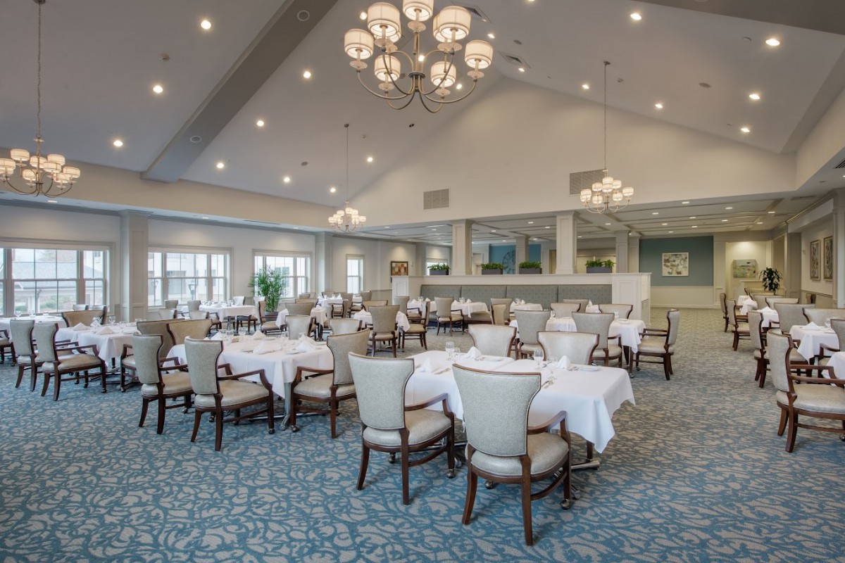 A spacious and elegant dining area featuring round tables with white tablecloths, upholstered chairs, and chandeliers hanging from the ceiling.