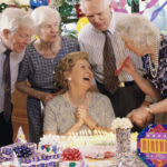 A joyful birthday celebration with a group of elderly friends gathered around a woman seated in front of a cake.