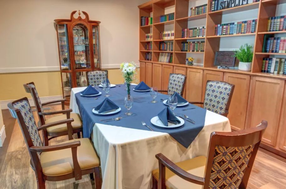 A neatly set dining table with blue napkins and a floral centerpiece, surrounded by wooden chairs and bookshelves.