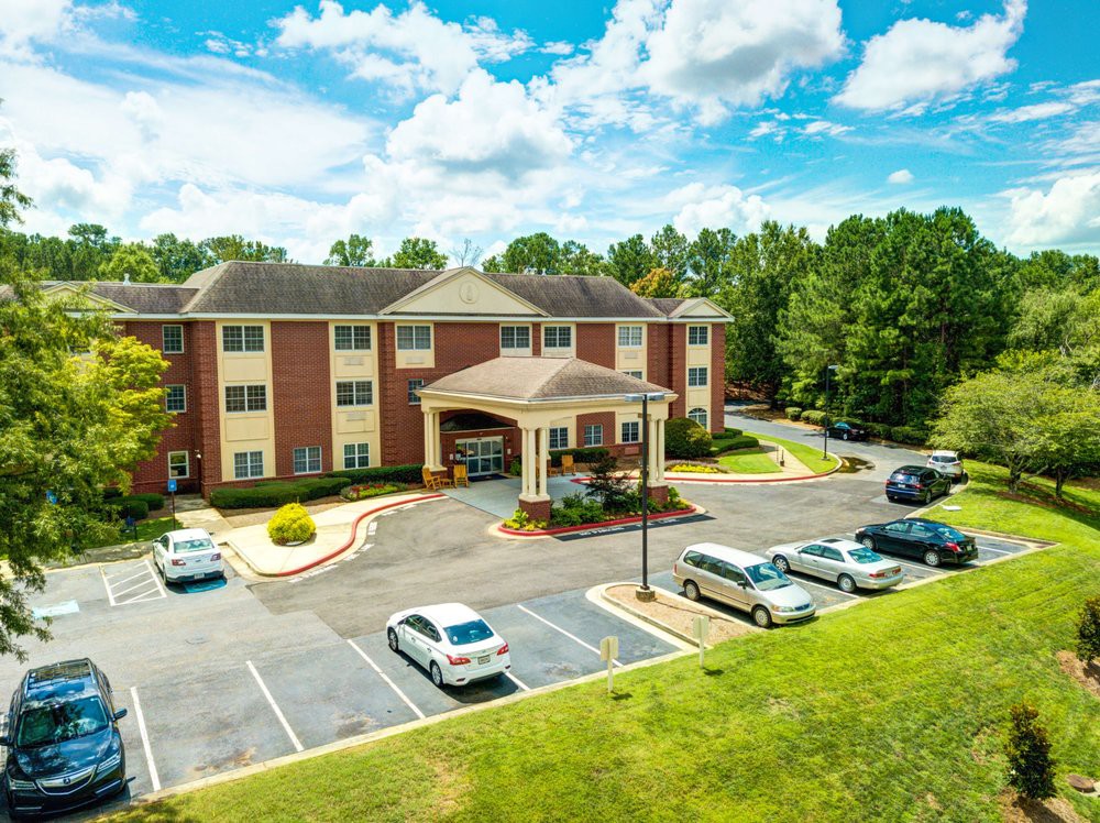 A three-story apartment building surrounded by a parking lot and lush trees under a partly cloudy sky.