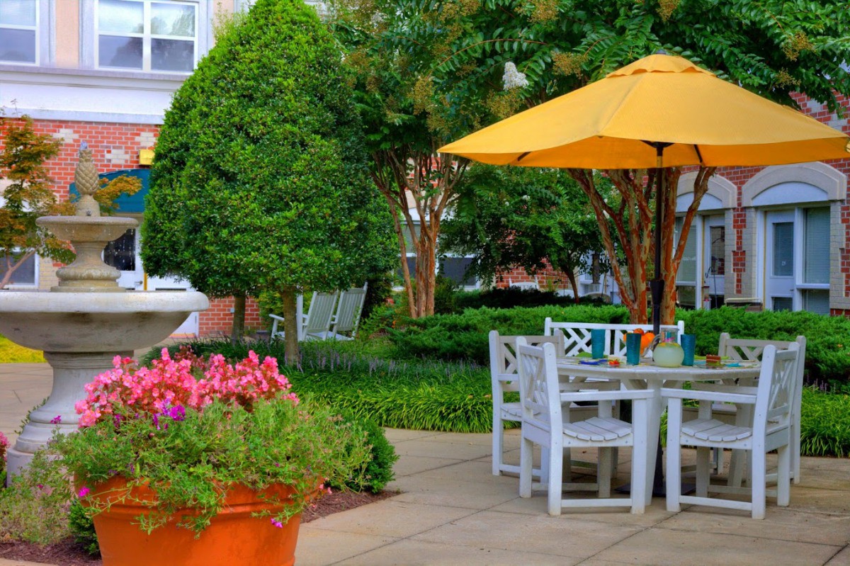 A peaceful patio setup with a table and chairs under a yellow umbrella, surrounded by lush greenery and colorful flowers.