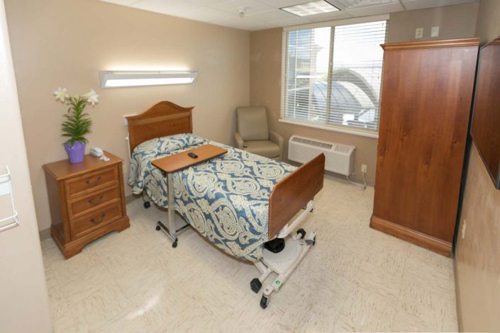 A clean and organized hospital room with a single bed, bedside cabinet, and chair.