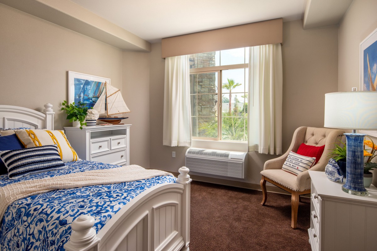A cozy bedroom with a nautical theme, featuring a bed, chair, dresser, and a large window with white curtains.