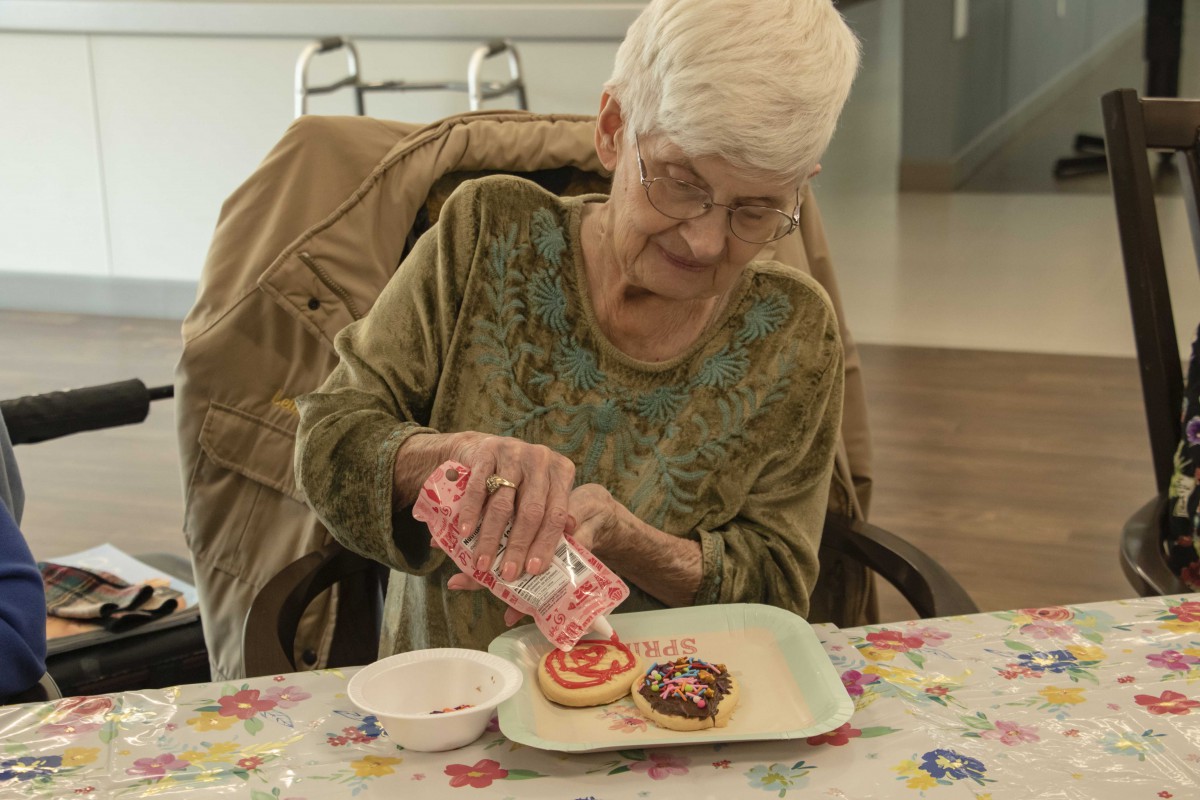 An elderly woman decorating cookies with icing and sprinkles at a table.