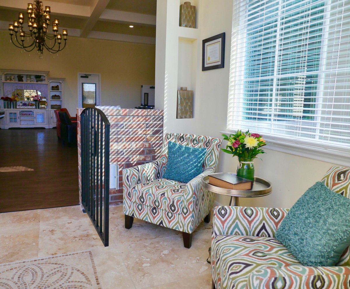A cozy living room featuring patterned armchairs, a small round coffee table with a vase of flowers, and natural light streaming through the window blinds.