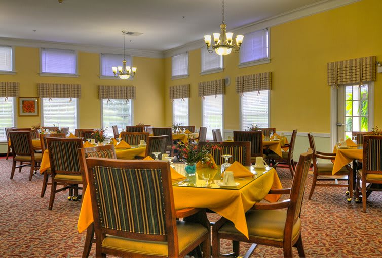 A well-lit dining room with neatly arranged tables and chairs, featuring yellow walls and chandeliers.