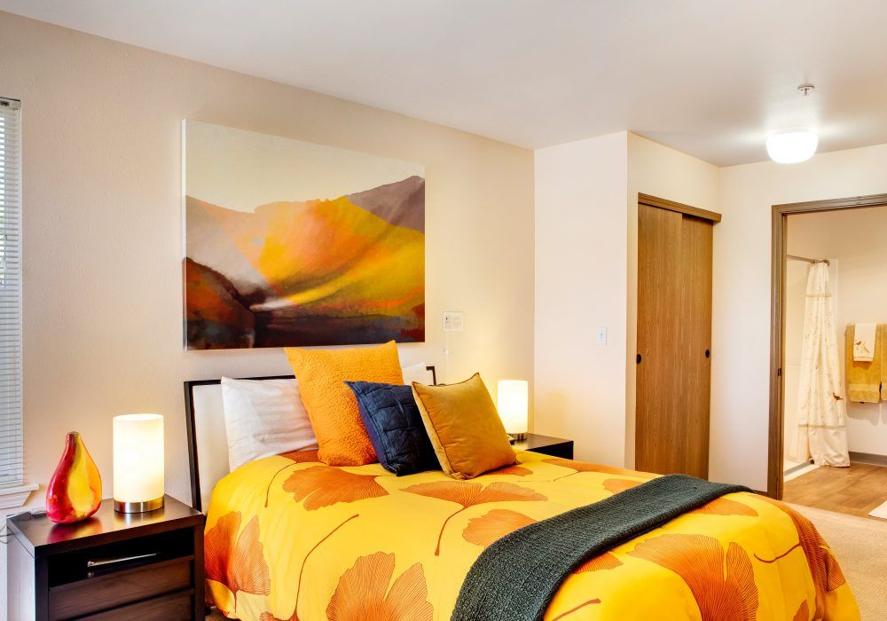 A modern bedroom featuring vibrant orange and yellow bedding, complemented by abstract wall art and warm lighting from bedside lamps.