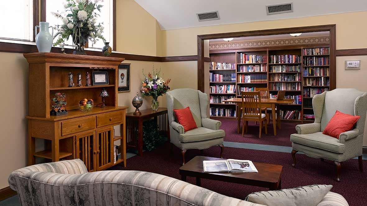 A cozy library reading room with an armchair, wooden table, and bookshelves filled with books.