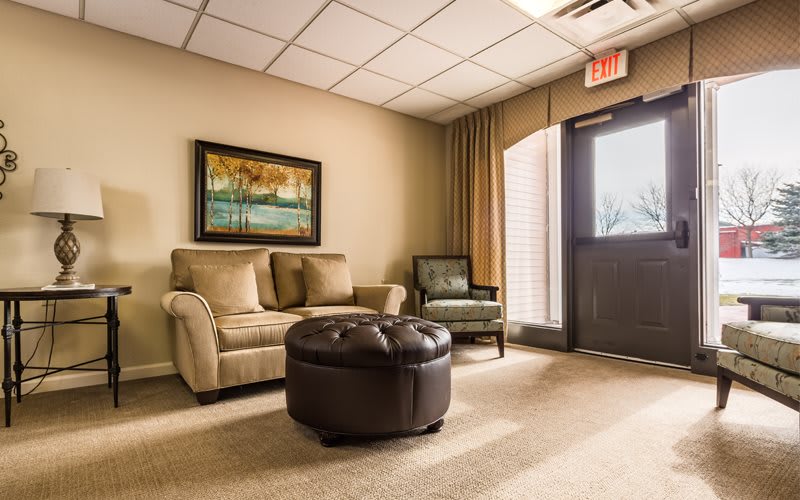 A cozy waiting room with a sofa, armchair, and ottoman, warmly lit by natural light from an open door.