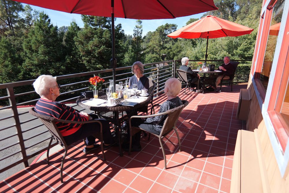 A group of seniors enjoying a meal on a sunny patio with red umbrellas and scenic views.