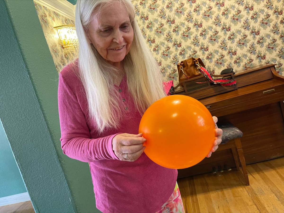 An elderly woman holding an orange balloon indoors.