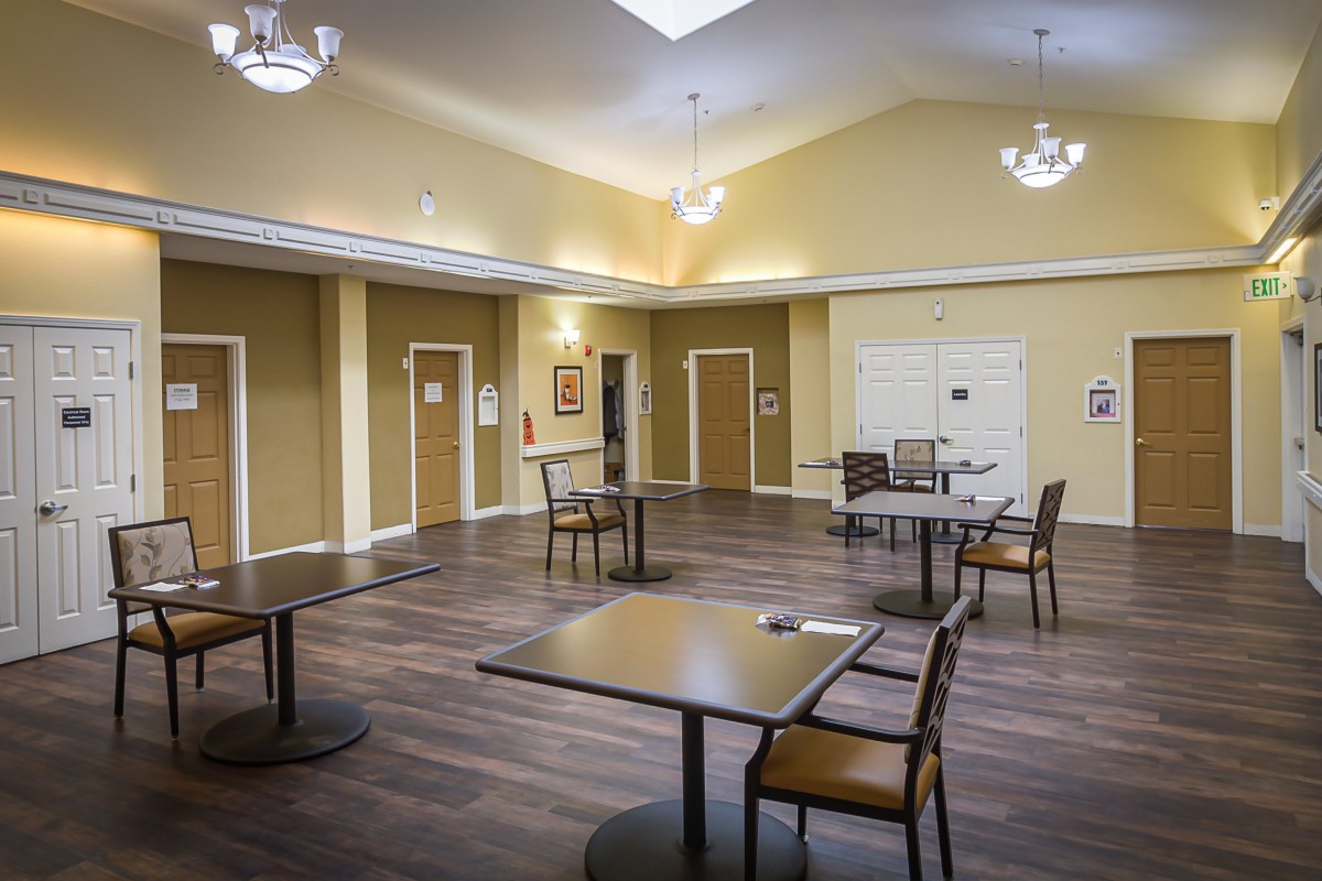 An empty room with several tables and chairs arranged on a wooden floor, illuminated by ceiling lights.