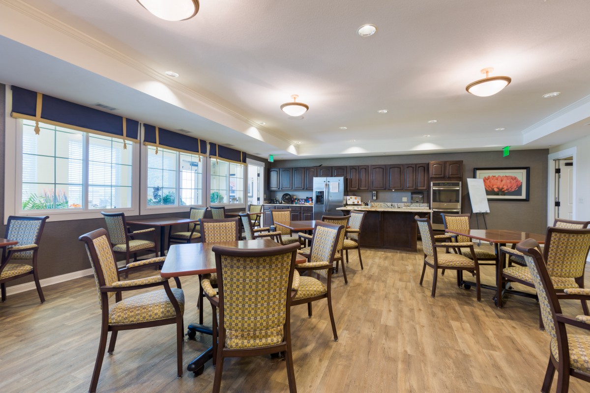A spacious dining and kitchen area with multiple tables and chairs, illuminated by ceiling lights and natural light from large windows.