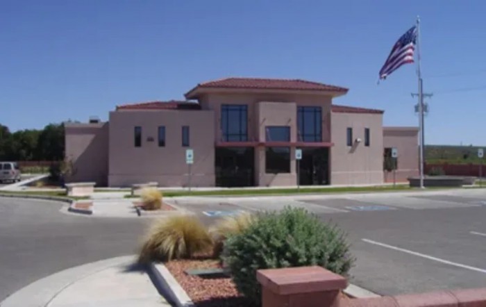 A modern building with an American flag in front, surrounded by a parking lot and desert landscaping on a sunny day.