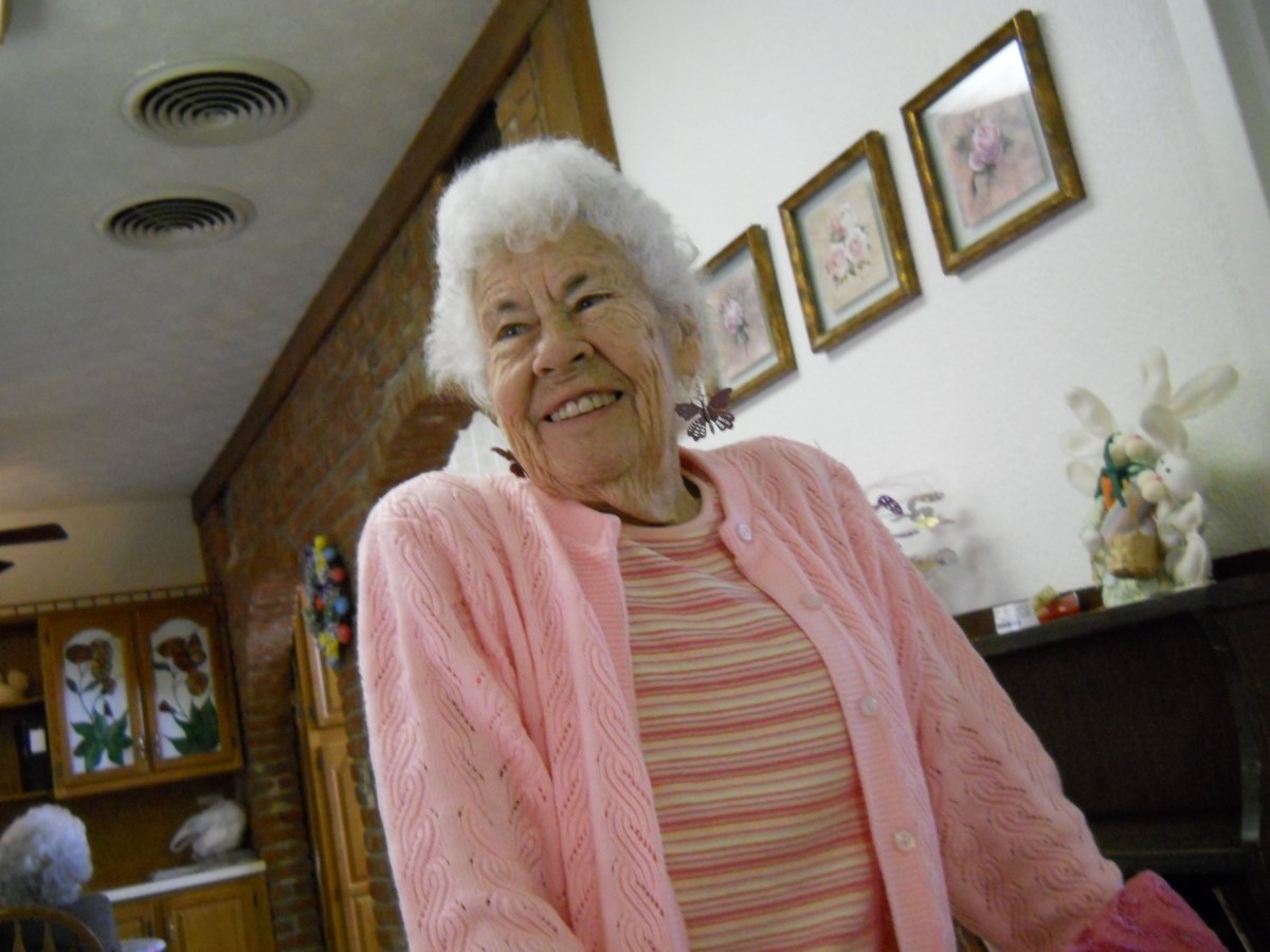 An elderly woman smiling warmly while wearing a pink sweater indoors.