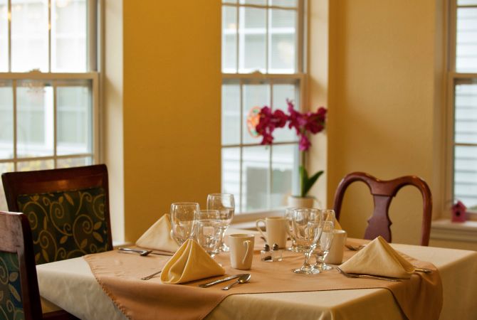 An elegantly set dining table with glasses, napkins, and a flower centerpiece by the window.