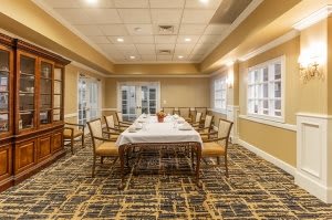 A well-lit dining room with a long table and chairs, featuring large windows and patterned carpet.