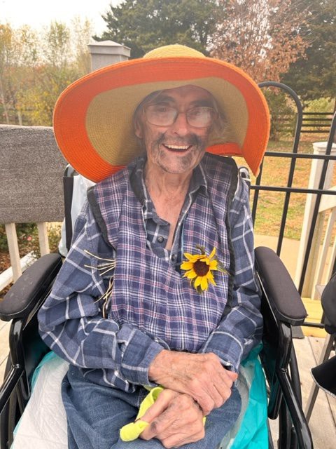 An elderly person sitting in a wheelchair, wearing a large hat and plaid shirt, with a sunflower attached to their clothing.