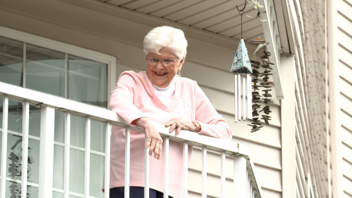 An elderly woman smiles gently as she looks down from a balcony, with a wind chime softly hanging nearby.