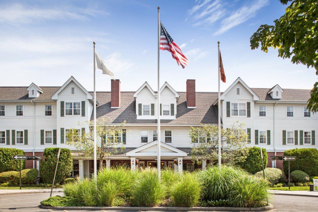A large colonial-style building with multiple flags on poles in front, surrounded by greenery and a clear sky above.