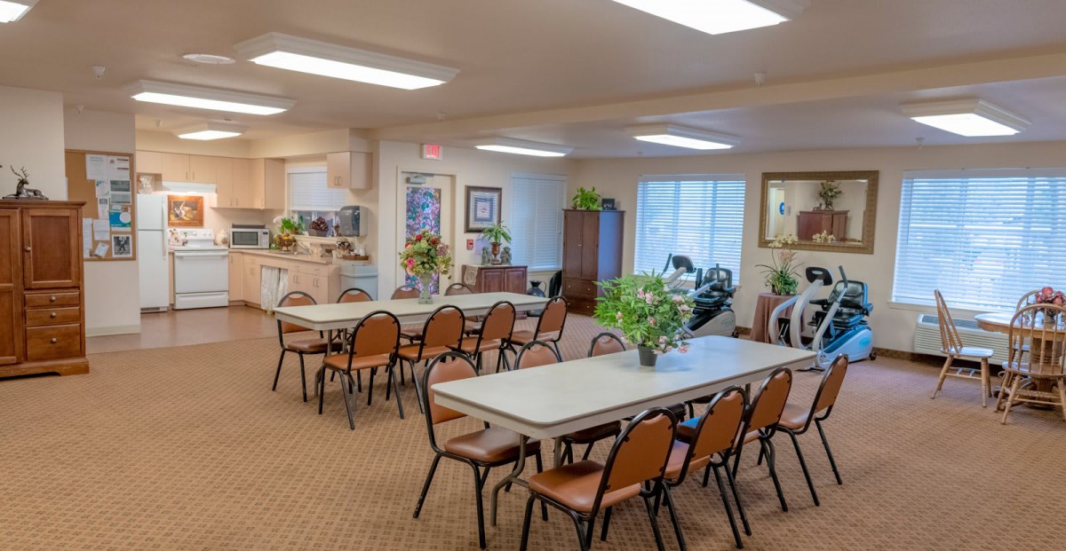A community dining area with tables and chairs, decorated with indoor plants, under bright lighting.
