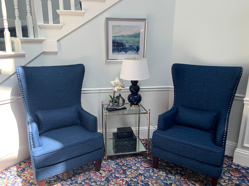 A cozy living room corner featuring two blue armchairs, a stylish side table with a lamp, and a piece of artwork on the wall.