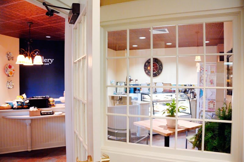A cozy cafe interior with a view through a window, featuring tables, plants, and a wall clock.