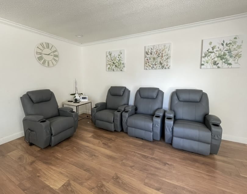A minimalist living room featuring four gray recliners, decorative wall art, a side table with a clock, and wooden flooring.