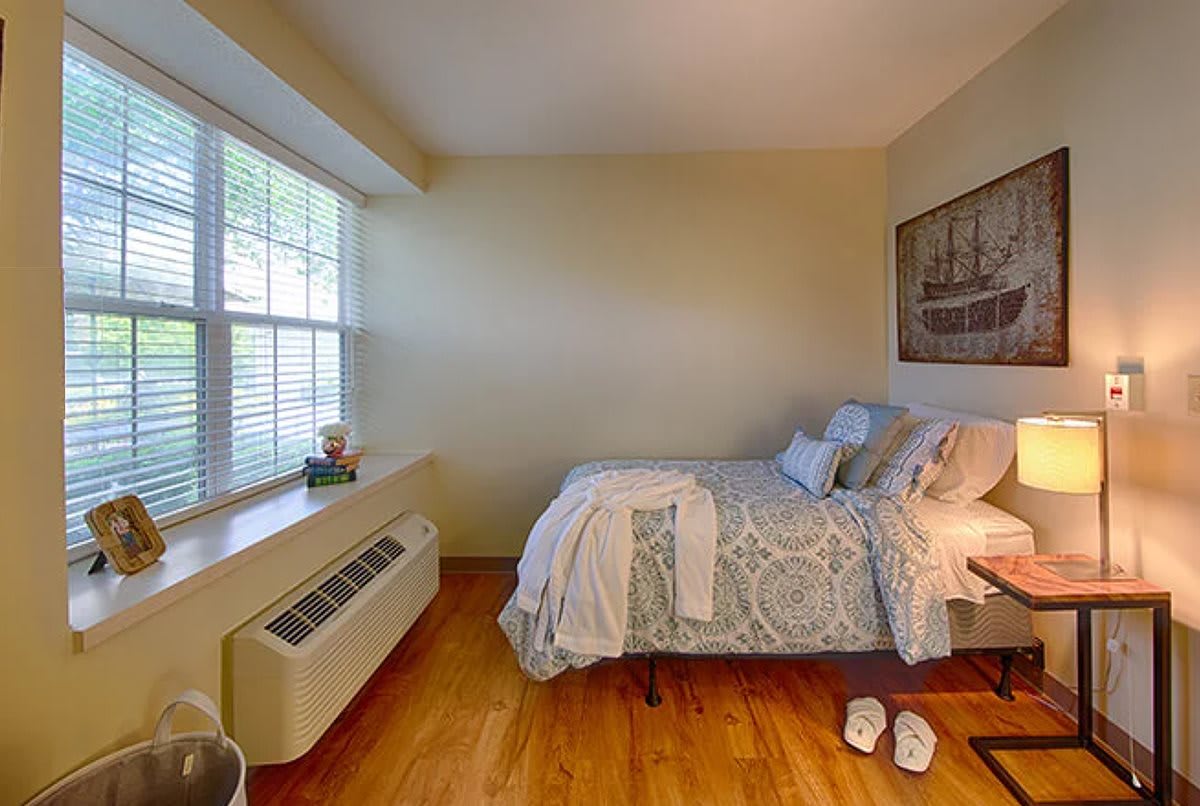 A cozy bedroom with a neatly made bed, wooden floors, and natural light coming through the window.