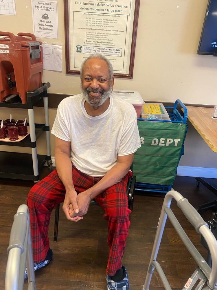 A man sitting indoors, smiling, wearing a white t-shirt and red plaid pants.