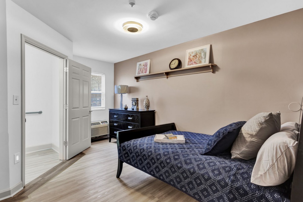 A cozy bedroom featuring a daybed with decorative pillows, a dresser with a lamp, and a wall shelf displaying artwork and collectibles.