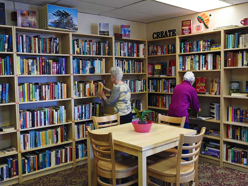 Two people browsing books in a cozy library with wooden shelves and a table in the foreground.