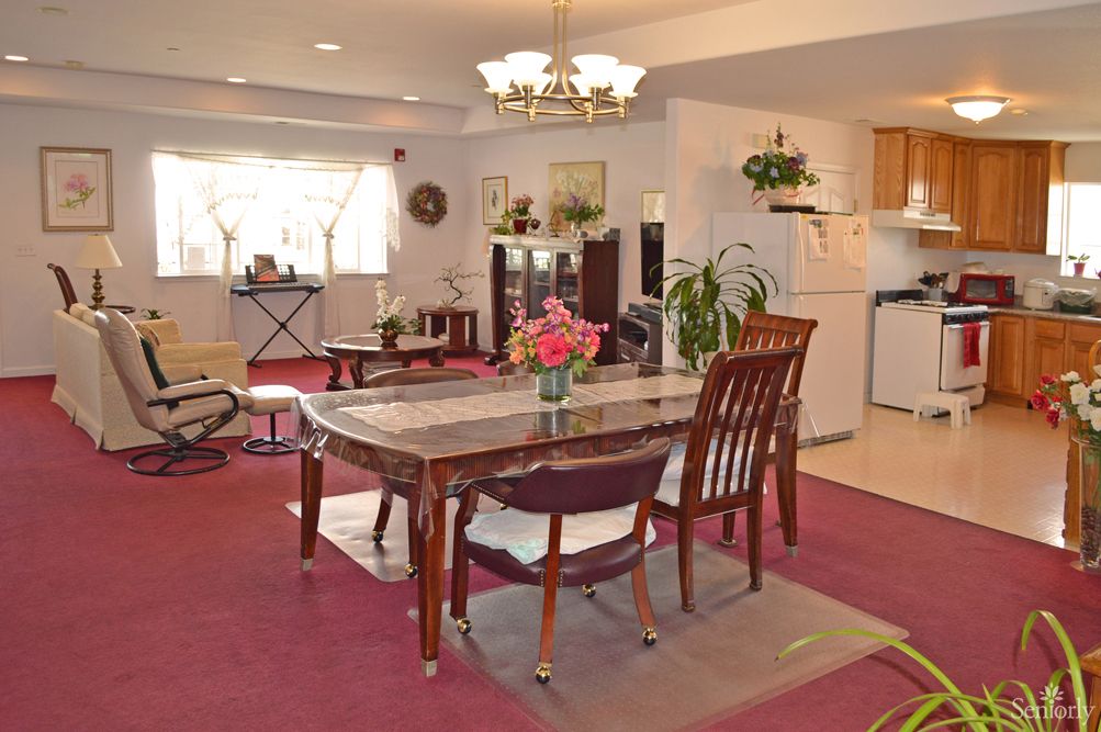 A spacious dining room with a table and chairs, decorated with plants and flowers, featuring a cozy carpet and adjacent open kitchen area.
