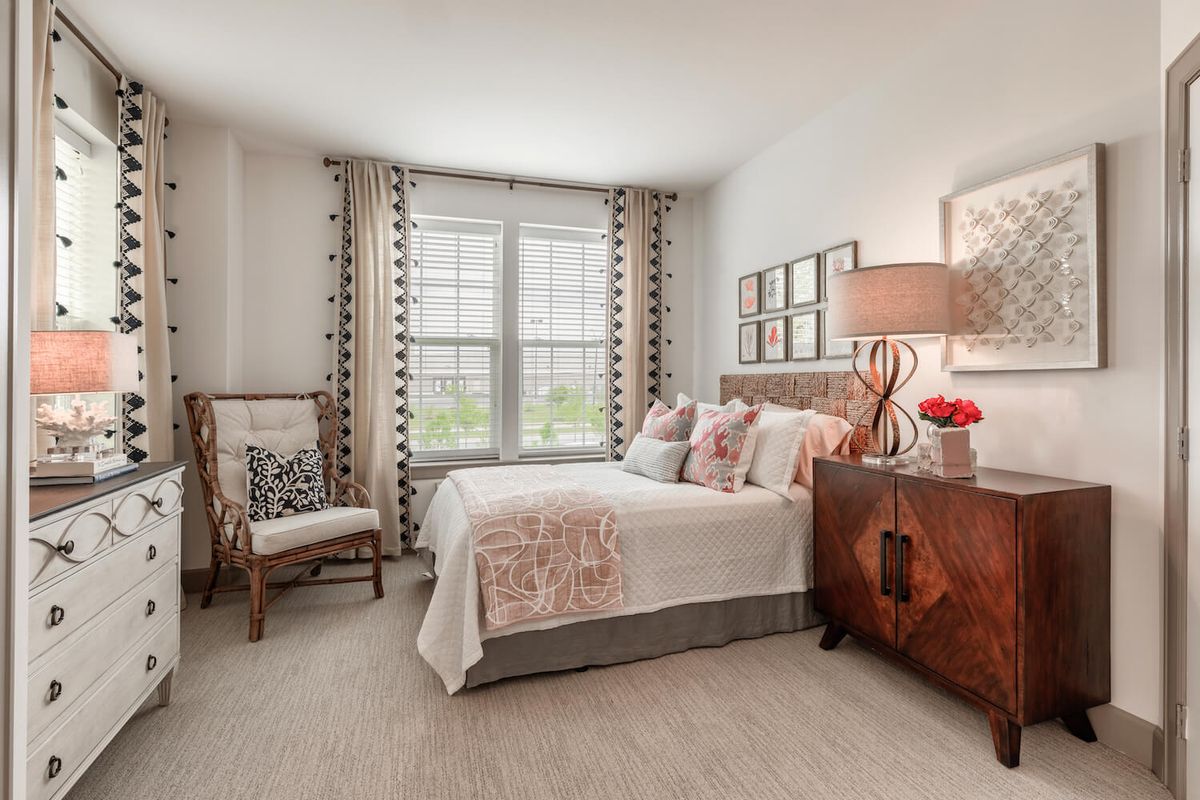 A modern bedroom with elegant decor, featuring a bed, chair, dresser, and ample natural light from large windows.