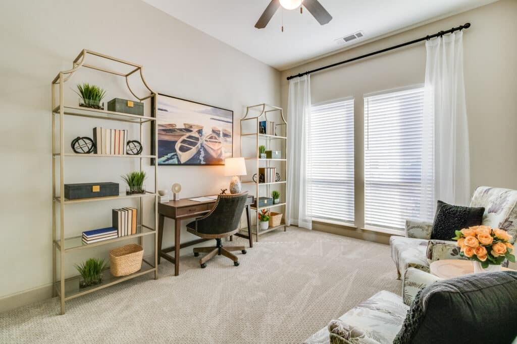 A modern home office with a desk, chair, and bookshelves, bathed in natural light from large windows.