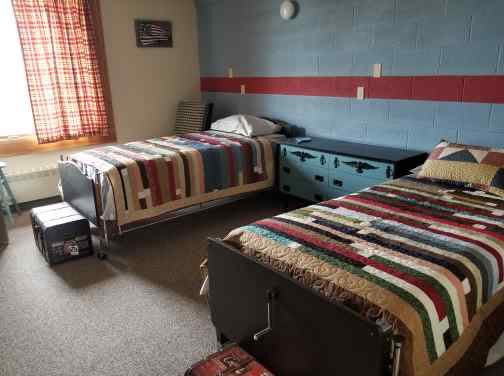 A cozy bedroom with two twin beds featuring colorful striped quilts and a window with curtains, creating a warm ambiance.