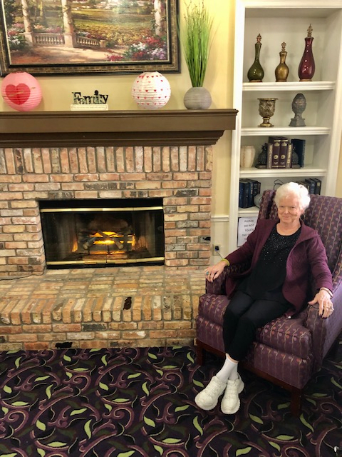 An elderly woman sitting in a chair next to a brick fireplace in a cozy living room.